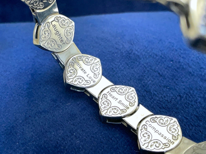 Close-up of engraved 14-karat white gold bracelet links with words like Laughter, Cultivate Love, Heart Smile, and Compassion on a royal blue background.