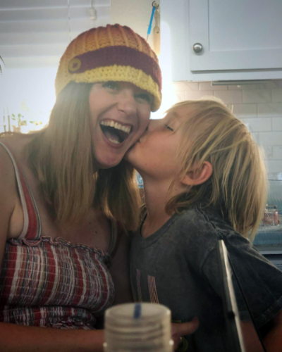 Woman and child sitting in a kitchen, child is kissing woman on the left cheek, while woman has laughter on her face and joy within her heart.