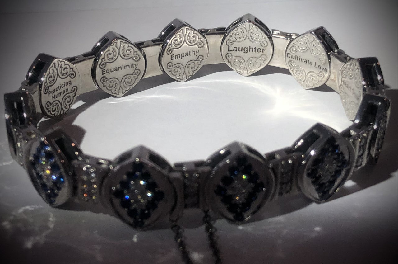 Close-up of engraved 14-karat white gold bracelet links with words like Laughter, Cultivate Love, Heart Smile, and Compassion on a royal blue background.